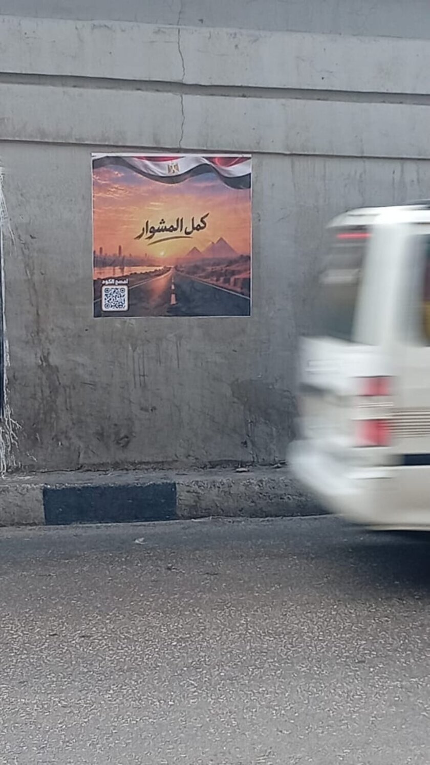 ملصقة كمل المشوار على حائط أسفل كوبري بينما تمر سيارة نقل بيضاء